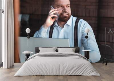 Young hipster, businessman talking over a phone in a cafe, laptop on the table Wall mural