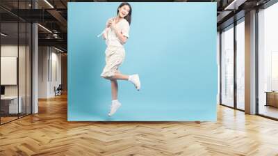 Beautiful young asian woman in white dress with flower pattern using smartphone and jump  isolated on blue background Wall mural