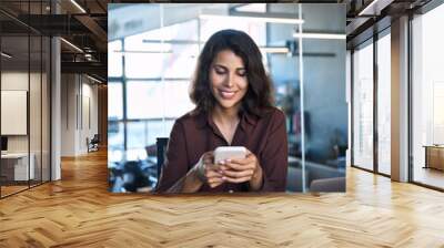 European business woman manager using cell phone mobile trading AI application. Smiling Latin Hispanic young female businesswoman entrepreneur holding smartphone sitting at desk in office. Vertical Wall mural