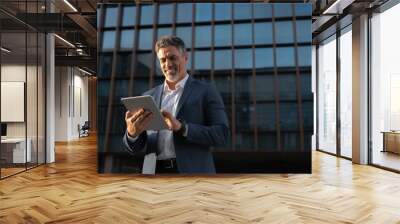 Focused successful mature Indian or Latin entrepreneur businessman holding digital pc tablet standing outdoor at business office building. Hispanic smiling man in suit working using touchpad computer Wall mural