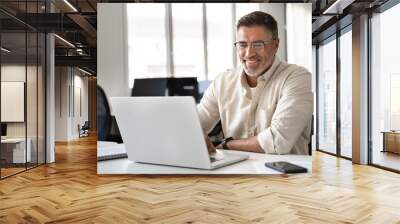 Happy smiling mature Indian or Latin business man ceo trader using computer, typing, working in modern office, doing online data market analysis, thinking planning tech strategy looking at laptop. Wall mural