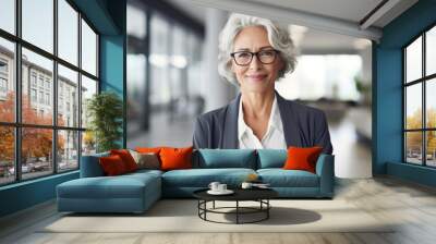 Mature business woman looking at camera indoors at work with copy space. Senior smiling European 50s 60s years businesswoman professional standing confident in modern coworking creative office space. Wall mural