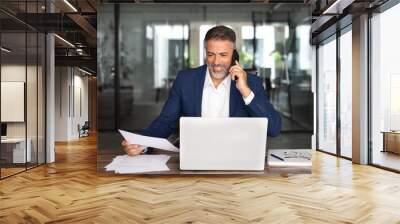 Middle aged Latin or Indian businessman having call on smartphone with business partners or clients. Smiling mature Hispanic man sitting at table talking by mobile cellphone and check data in office. Wall mural