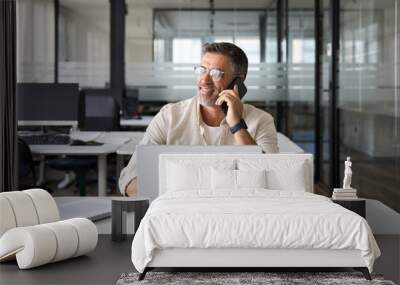 Middle aged Latin or Indian businessman having call on smartphone with business partners or clients. Smiling mature Hispanic man sitting at table talking by mobile cellphone at workplace in office.  Wall mural