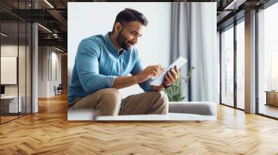 Millennial indian man using tablet relaxing on couch sofa in living room Wall mural