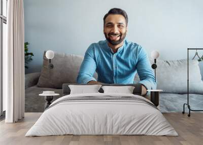 Portrait of indian man toothy smiling to camera sitting on sofa webcam view Wall mural