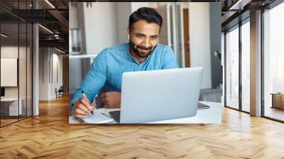 Portrait of mature adult indian man wearing earphones looking at laptop screen. Motivated student writing notes during online lesson, watching webinar, learning language online, sitting at work desk Wall mural