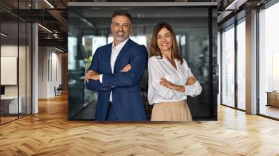 Portrait of smiling mature Latin or Indian business man and European business woman standing arms crossed in office. Two diverse colleagues, group team of confident professional business people. Wall mural