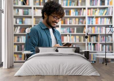 Side view portrait of happy young Indian student chatting by mobile phone in university library. Teen guy wearing denim shirt and eyeglasses using social media application browsing on smartphone Wall mural