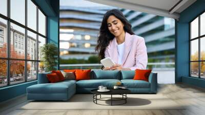 Smiling young middle eastern Israel businesswoman using smartphone mobile phone online app for work at office building. Successful 20s happy indian or arabic woman in business suit holding cellphone Wall mural