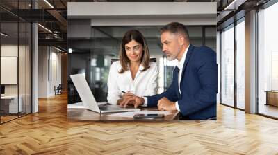 Team of diverse partners sitting at table mature Latin business man and European business woman discussing project on laptop in office. Two colleagues of professional business people working, close up Wall mural