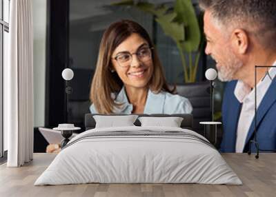 Vertical shot of team of diverse partners mature Latin business man and European business woman discussing project at table in office. Two colleagues of professional business people working on tablet. Wall mural