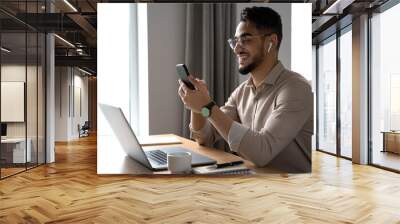 Young adult Arabic businessman holding cellphone smartphone at hands, sitting at desk with laptop computer at home office. Smiling Indian man using phone application, pc for business freelance work Wall mural