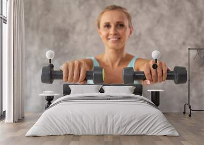 Smiling woman in sport outfit holds two dumbbells in hands. Portrait photo. Selective focus Wall mural