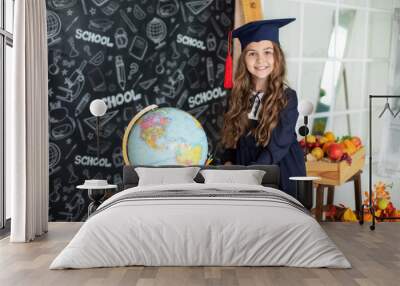 School Kid in graduate headdress is studying in classroom against of blackboard. Smiling little girl in a school uniform and master's hat holding globe in classroom. Back to school, learning lessons.  Wall mural