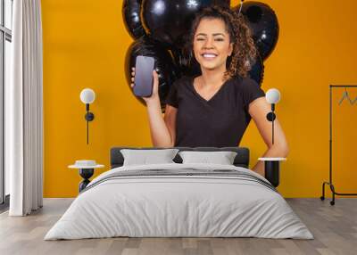 Smiling charming young afro woman holding a cellphone with empty blank screen on yellow background with air balloons studio portrait. Black Friday settlement Wall mural