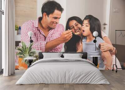 An attractive happy, smiling Asian Indian family of mother, father and daughter. Father feeding cereal to daughter with spoon at dining table. Indians eating breakfast, lunch / dinner. Selective focus Wall mural