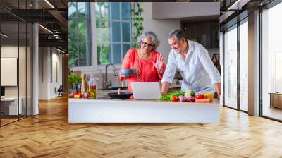 Indian asian senior old age cheerful couple using tablet computer or laptop in the kitchen while cooking Wall mural