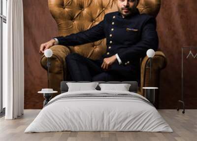 Indian bridegroom wears ethnic or traditional cloths,  Male fashion model with dark blue sherwani, Sitting on Wing Chair / sofa over brown grunge background, selective focus Wall mural