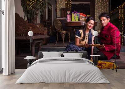 Indian young couple in traditional wear lighting oil lamp and celebrating Diwali or deepavali, fesitval of lights at home. Husband holding a plate full of oil lamp indoors near flower rangoli Wall mural