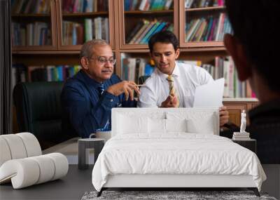 Two Indian senior and young businessmen discussing at the table together with laptop and paper, showing library in the background Wall mural