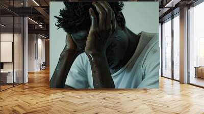 A distressed young black man with short dark hair indoors wearing a white t shirt sits with head in hands looking down in an image with copy space Wall mural