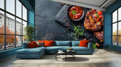 Freshly grilled pork chops, steaks, vegetables, coriander, beans, and sauce displayed on a black slate with a dark background, accented with herbs and foam, creating an artistic copy space image. Wall mural