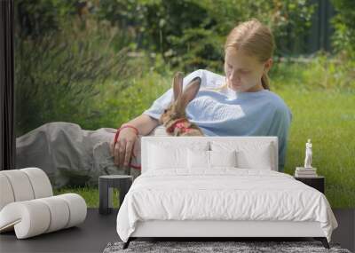 A young woman lies on the grass with a rabbit in a red harness, enjoying a relaxed outdoor moment together. Wall mural