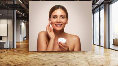 Woman applying facial moisturizer against a beige background Wall mural