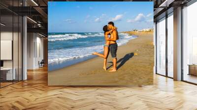 Happy young couple in bikini and shorts enjoying summer at the beach at dusk, breathing fresh air, kissing and teasing one another Wall mural