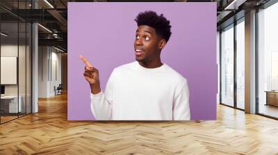 Happy funny African American teen guy pointing fingers aside advertising new promo offer. Smiling black student model showing presenting ads standing isolated on purple background Wall mural