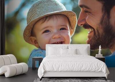 A cheerful baby wearing a hat is held by a smiling father outdoors on a sunny day, both radiating happiness and warmth. Wall mural