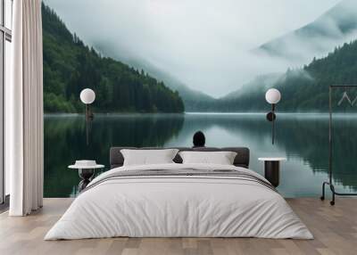 a man stands at the edge of a calm lake and looks to the mountains Wall mural