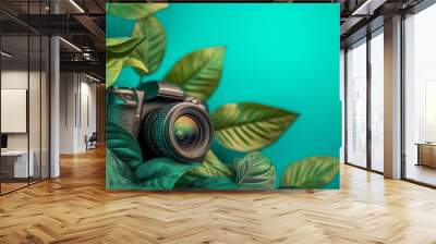 Close-up of a digital camera amidst vibrant green leaves, set against a teal background, representing photography and nature. Wall mural