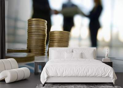 Multiple stacks of coins on a desk, with blurred figures in a business meeting handshake in the background. Wall mural