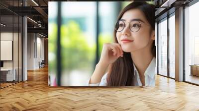 Thoughtful young woman with glasses looking out of the window, sitting at a table in a modern cafe, surrounded by green plants. Wall mural