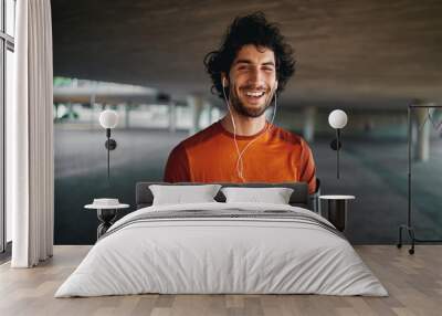 Cheerful portrait of a healthy sporty young man enjoying listening to music on earphones standing on city street Wall mural