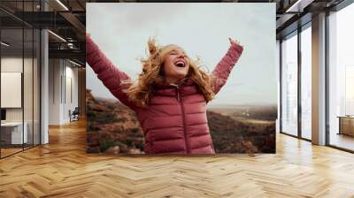 Cheerful young woman with hands raised and outstretched mountaineer with hair flying in wind enjoying fresh breeze - happiness and travel - achieving your dreams Wall mural