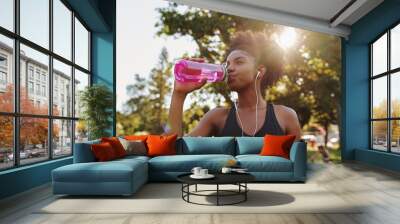 Fitness athlete young african american woman listening to music on earphones drinking water in a reusable water bottle after working out exercising on sunny day at the park Wall mural