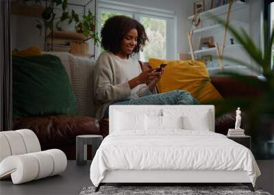 Happy young african woman using mobile phone while sitting a couch at home Wall mural