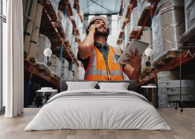 Low angle view of warehouse manager talking over phone while smiling and holding digital tablet standing in aisle with goods Wall mural