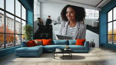 Portrait of a friendly successful young black businesswoman holding digital tablet in hand looking away at office Wall mural