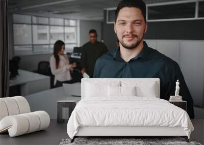 Portrait of a professional young businessman smiling and looking at camera while other business people talking in background at office Wall mural