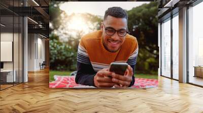 Portrait of an african smiling young man student wearing black eyeglasses relaxing in park using smart phone - college student using mobile in park Wall mural