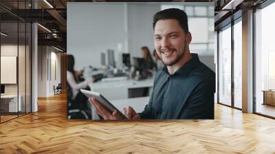 Portrait of confident smiling young businessman holding digital tablet in hand with his colleagues working at desk in background Wall mural