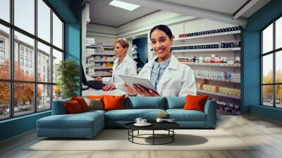 Portrait of smiling young female pharmacist wearing labcoat holding digital tablet with senior colleague working in background on computer Wall mural