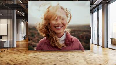 Portrait of young smiling woman face partially covered with flying hair in windy day standing at mountain - carefree woman Wall mural