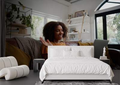 Smiling young african american woman relaxing on the couch waving hand while talking to friends on a video call at home - young blogger working from home Wall mural