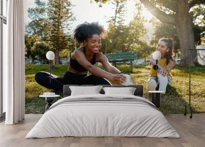 Young diverse female friends sitting on green grass stretching her legs in the morning sunlight at park - diverse friends warming up before doing group exercise  Wall mural