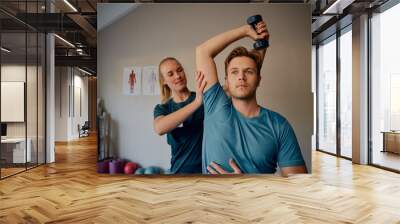 Young female physiotherapist helping patient to raise dumbbells in rehabilitation centre Wall mural